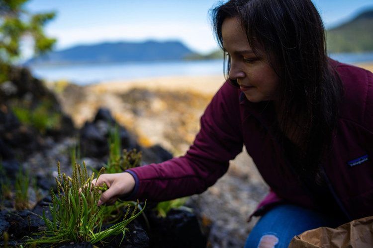 A woman is kneeling down on the beach looking at a plant.