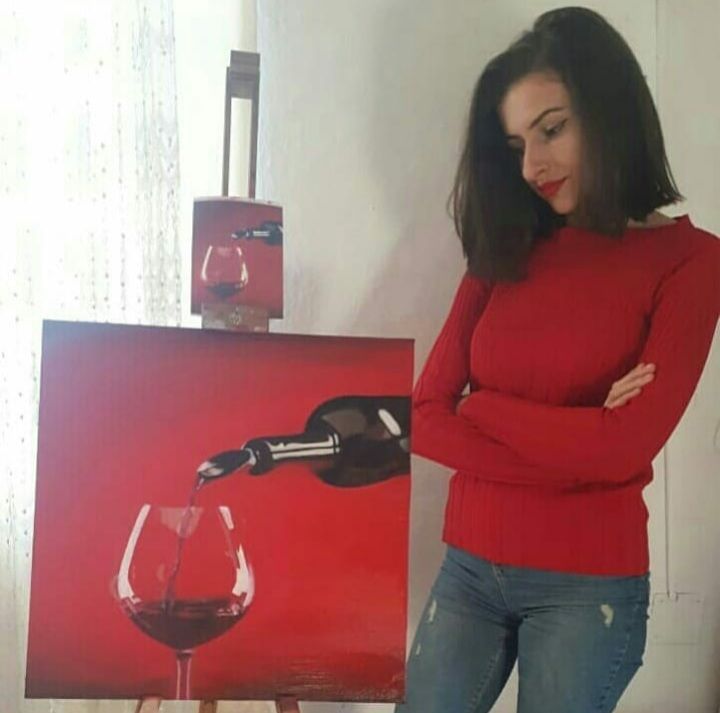 A woman in a red sweater is standing next to a painting of a bottle pouring wine into a glass