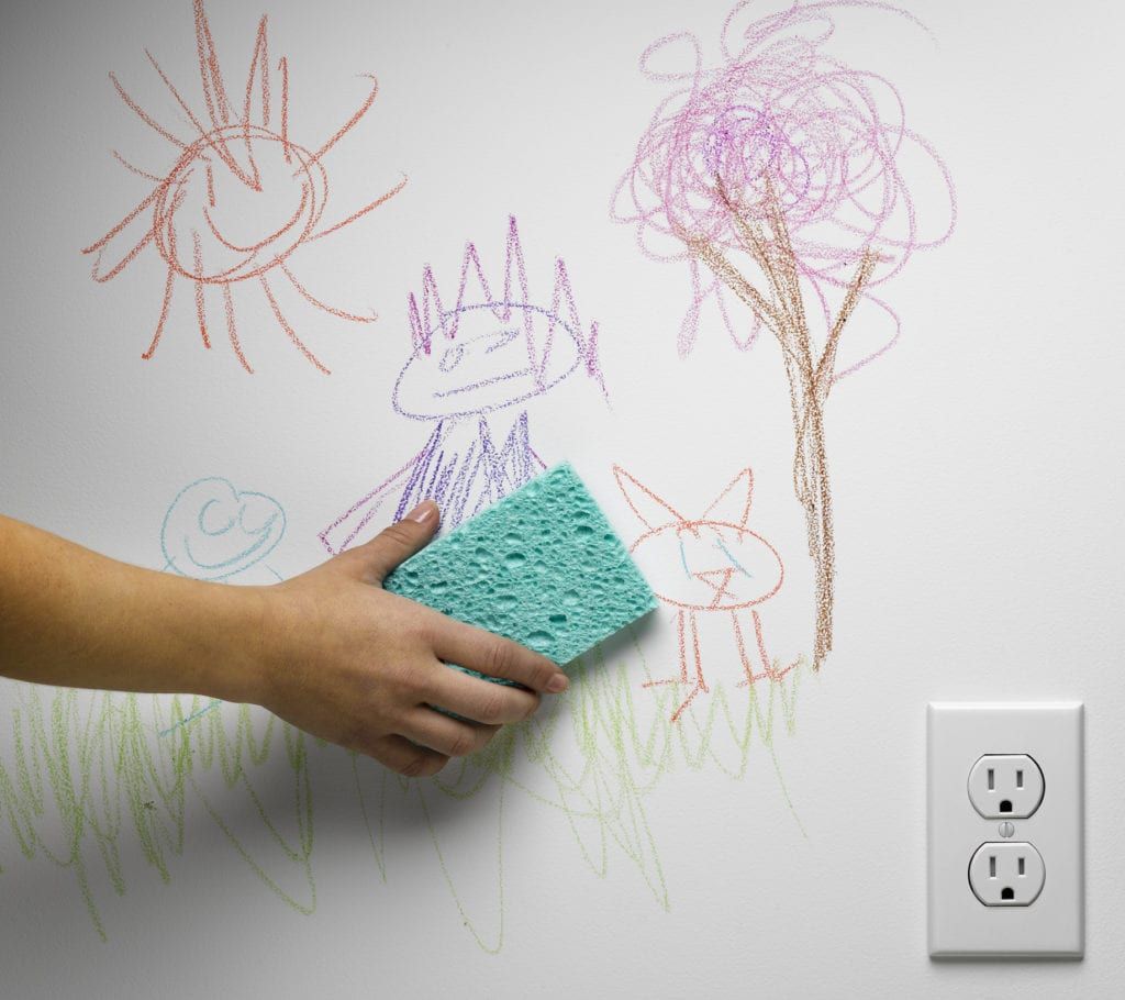 Hand with sponge erasing crayon drawings of a sun, tree, and cat from a white wall next to an electrical outlet.