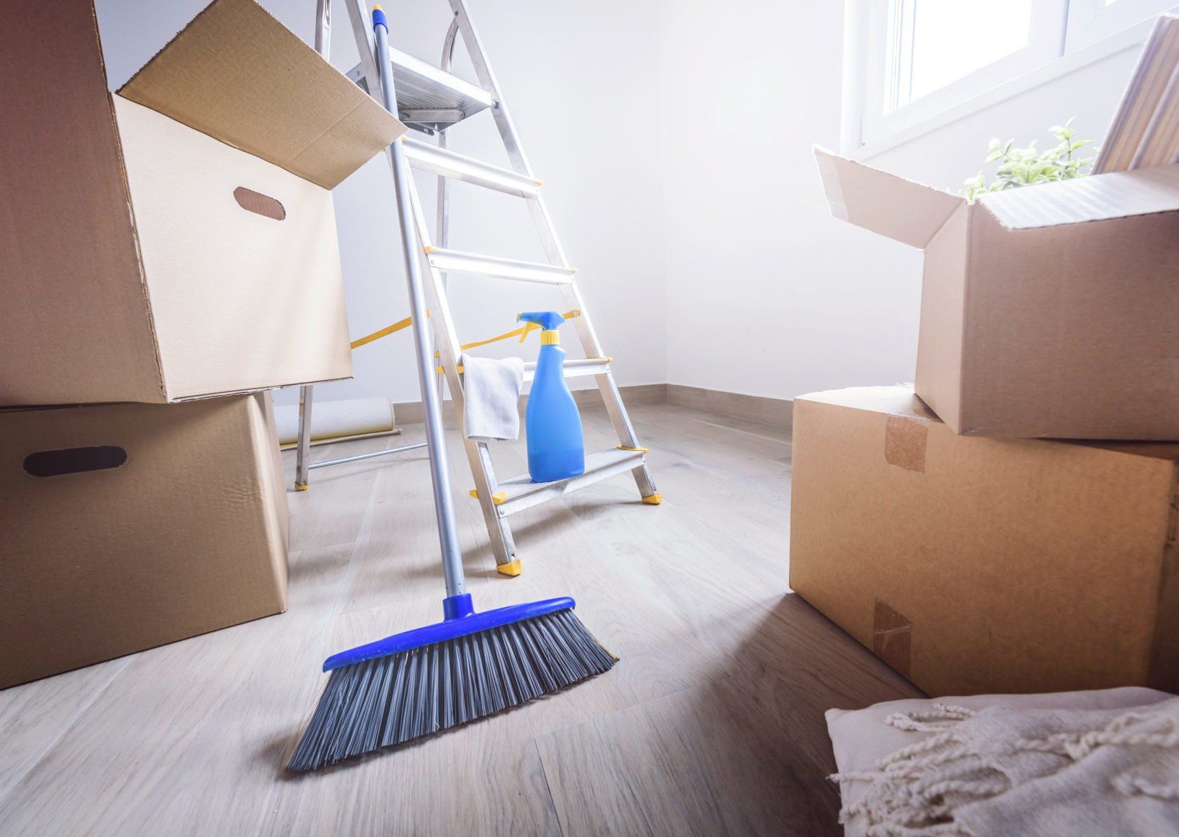Boxes, ladder, broom, and spray bottle in a room, indicating moving or cleaning.