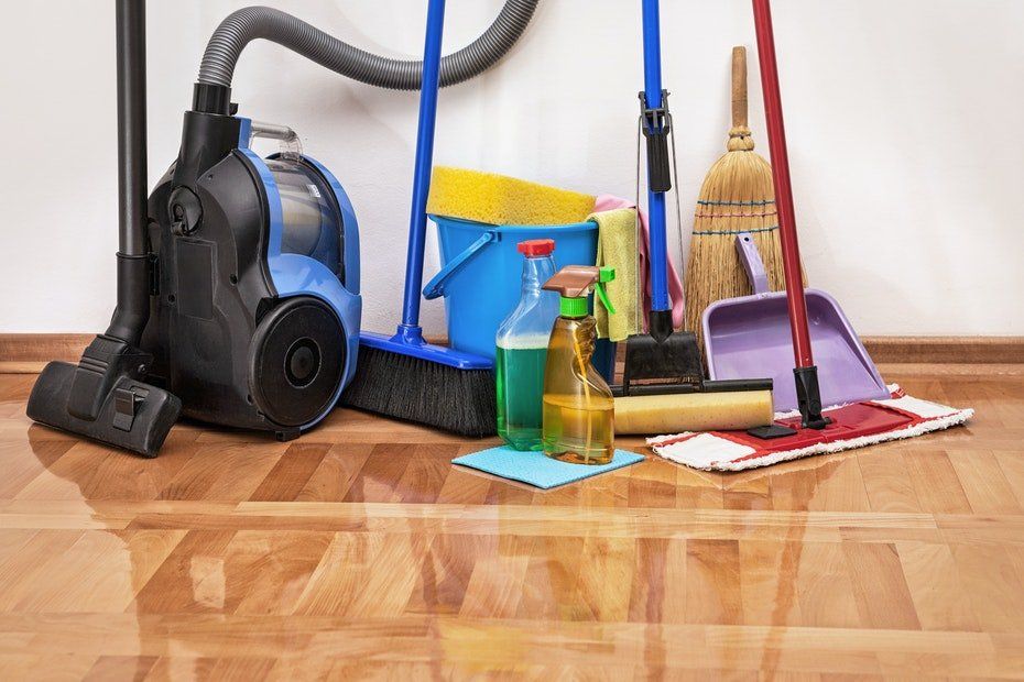 Cleaning supplies on a wooden floor: vacuum, brooms, bucket, spray bottles, mop, dustpan.