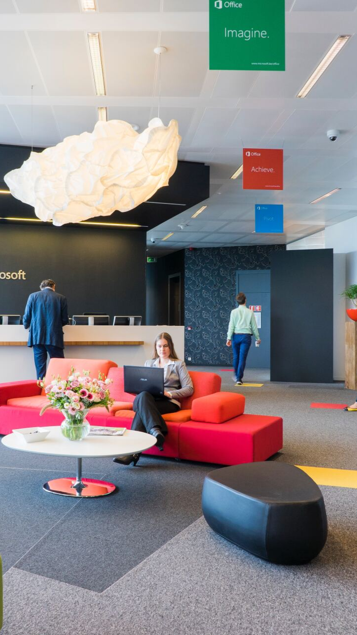Modern office lobby with red and orange seating, a woman using a laptop, and Microsoft signage.
