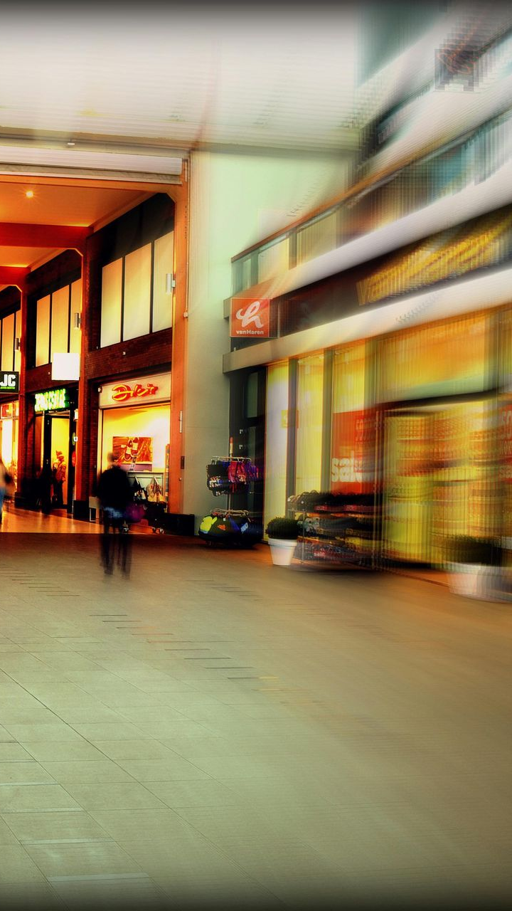 Blurred photo of a shopping mall interior with shops, people, and warm lighting.