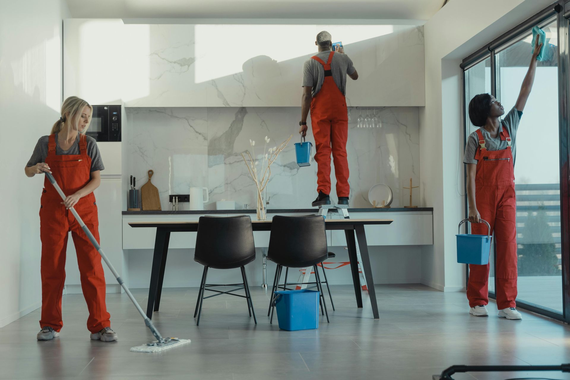 Three people in orange jumpsuits cleaning a modern kitchen. One mops, one paints, and one wipes a window.