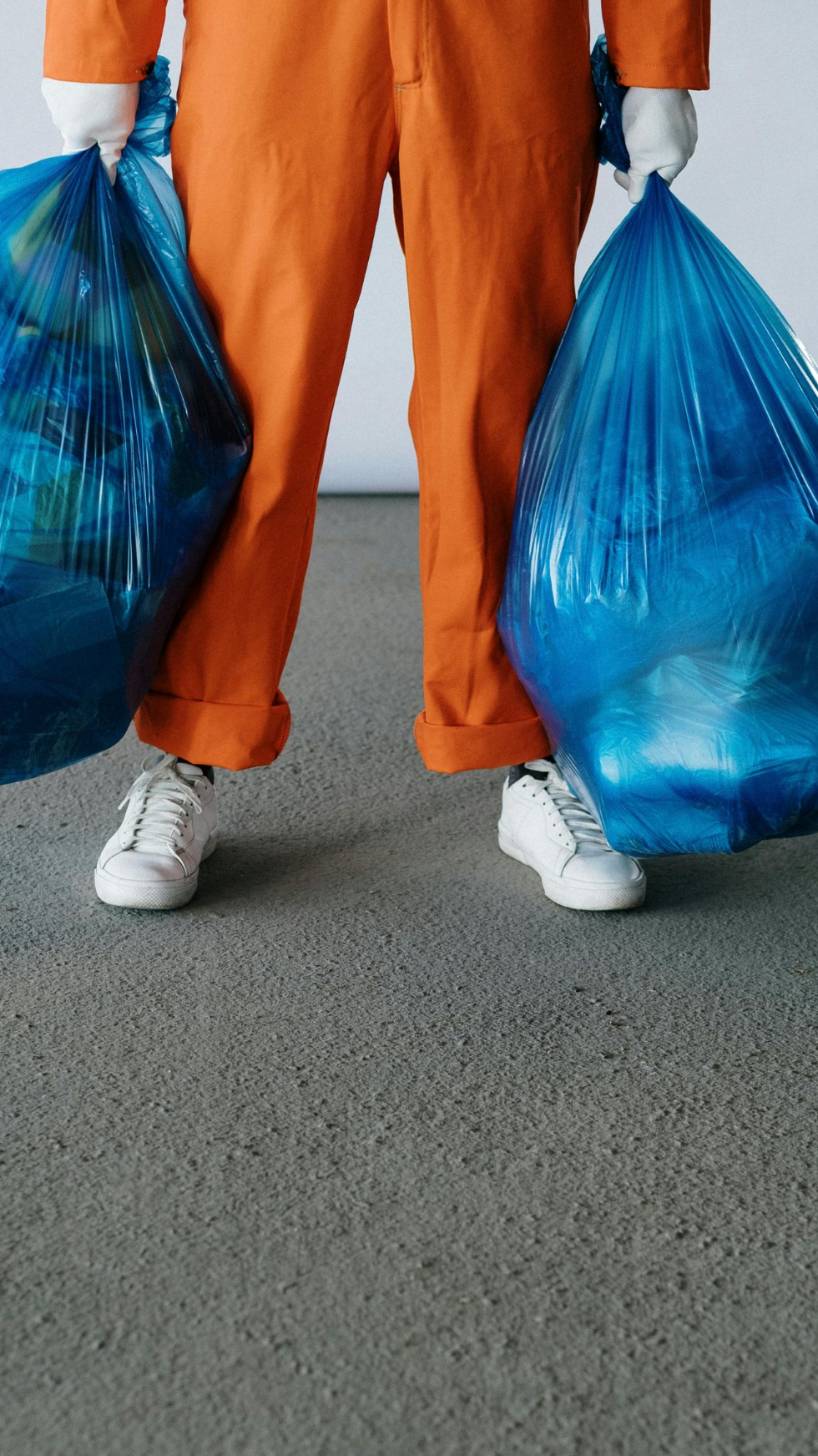 Person in orange jumpsuit and white gloves holds two full blue trash bags.