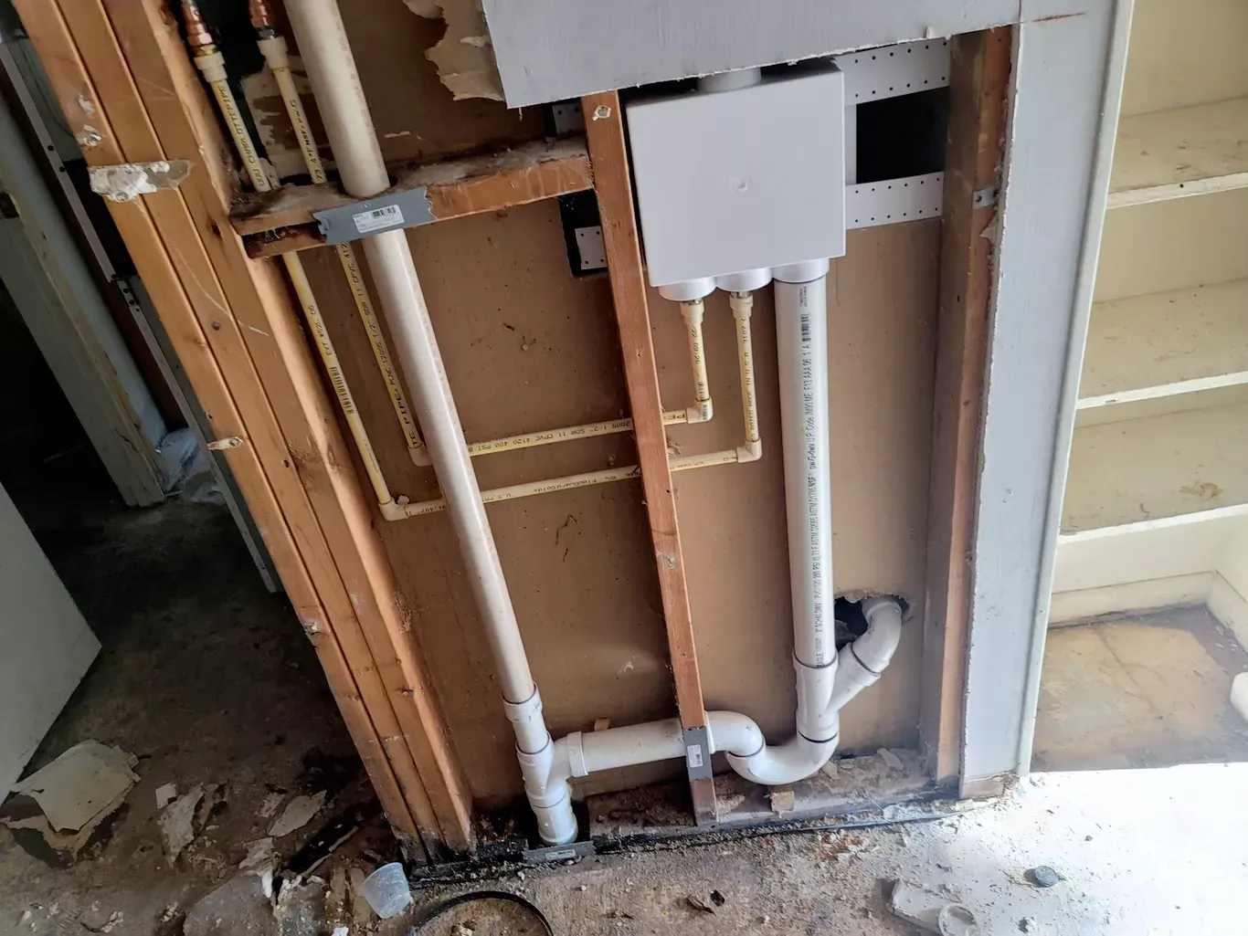 Plumbing rough-in for two toilets inside a wall framed with metal studs; a ladder leans nearby.