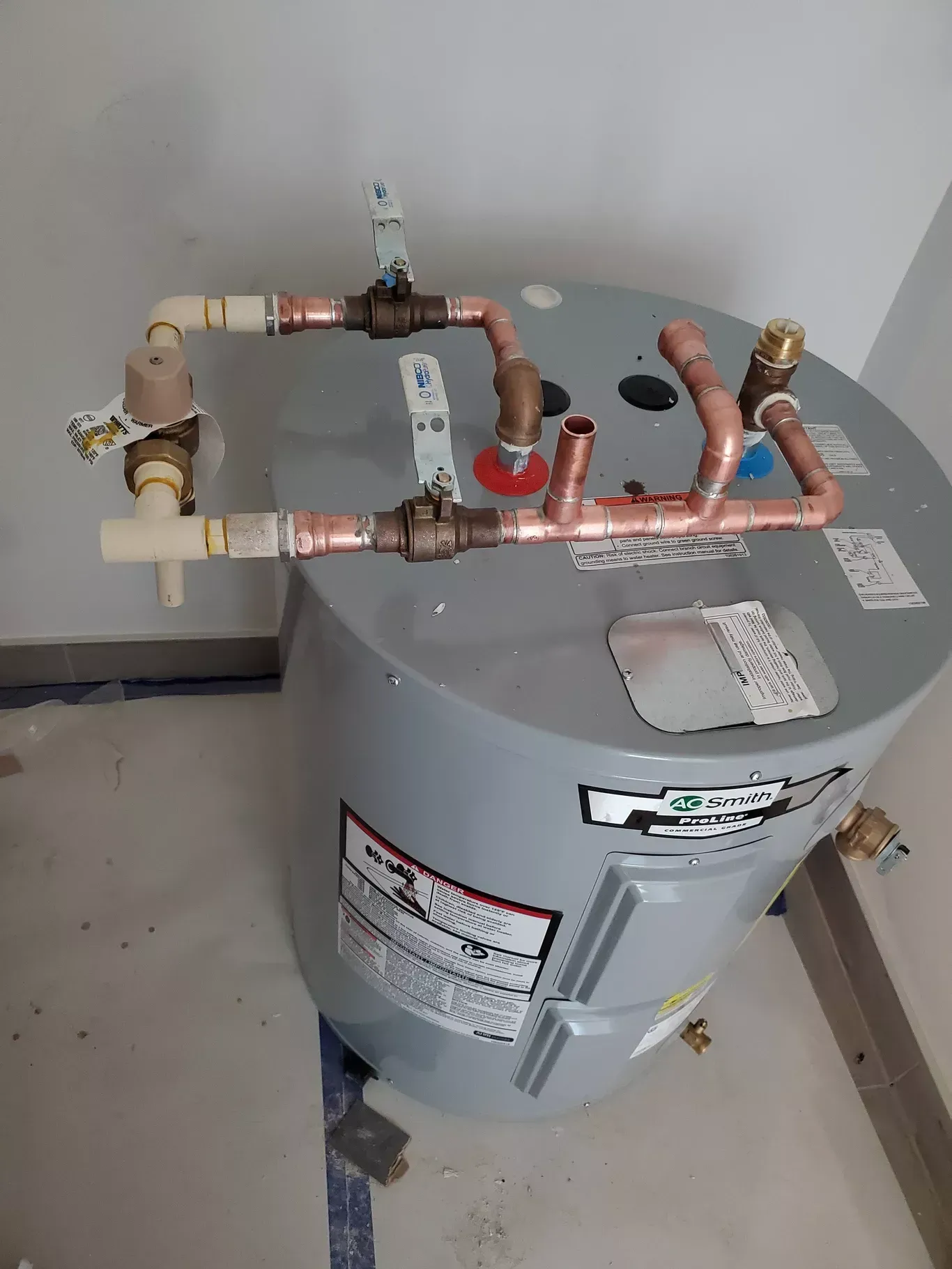 A water heater with copper pipes attached to it is sitting on the floor.