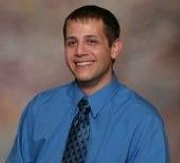Man in blue shirt and patterned tie smiles; blurred background.
