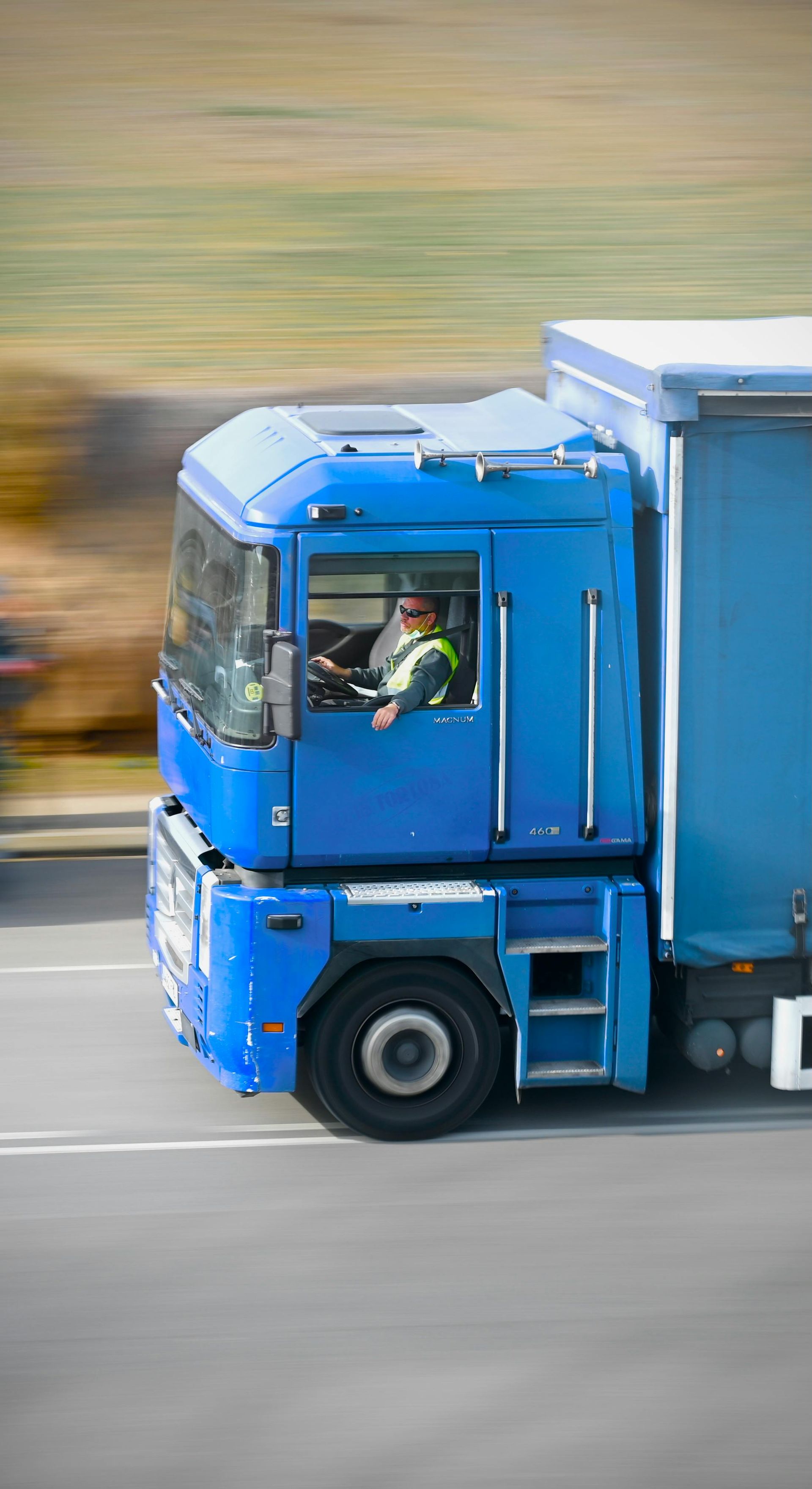 Blue semi-truck speeding on a highway, driver visible in the cab, blurred background.