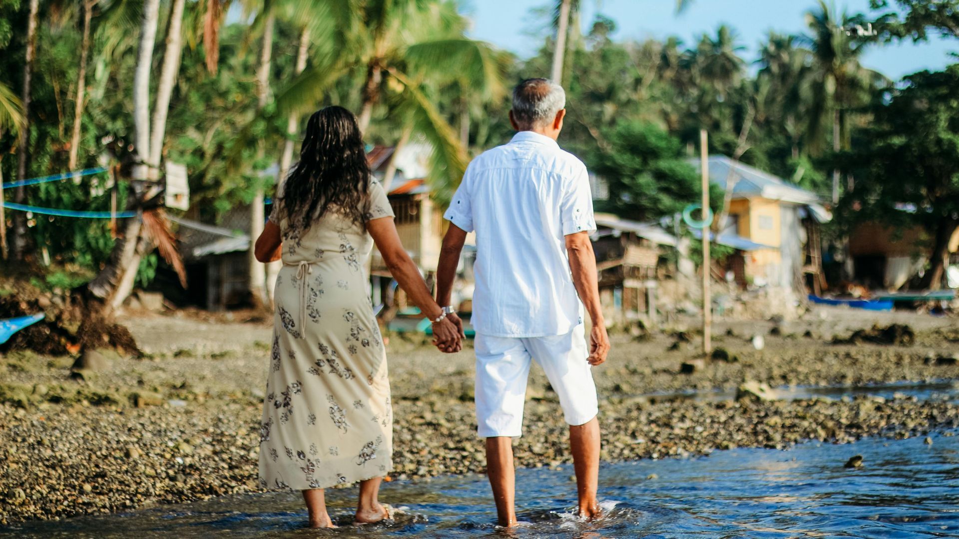 Couple holding hands, wading into water, looking at the distant shore. Green palm trees and houses in background.