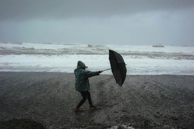 Person struggles with umbrella on a stormy beach. Dark sky, waves crash, person in jacket.