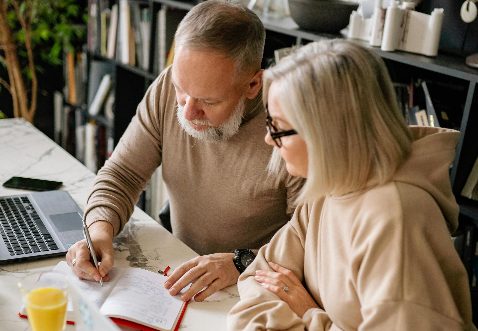An older couple reviews paperwork at a table with a laptop, books, and a glass of juice; the man writes.