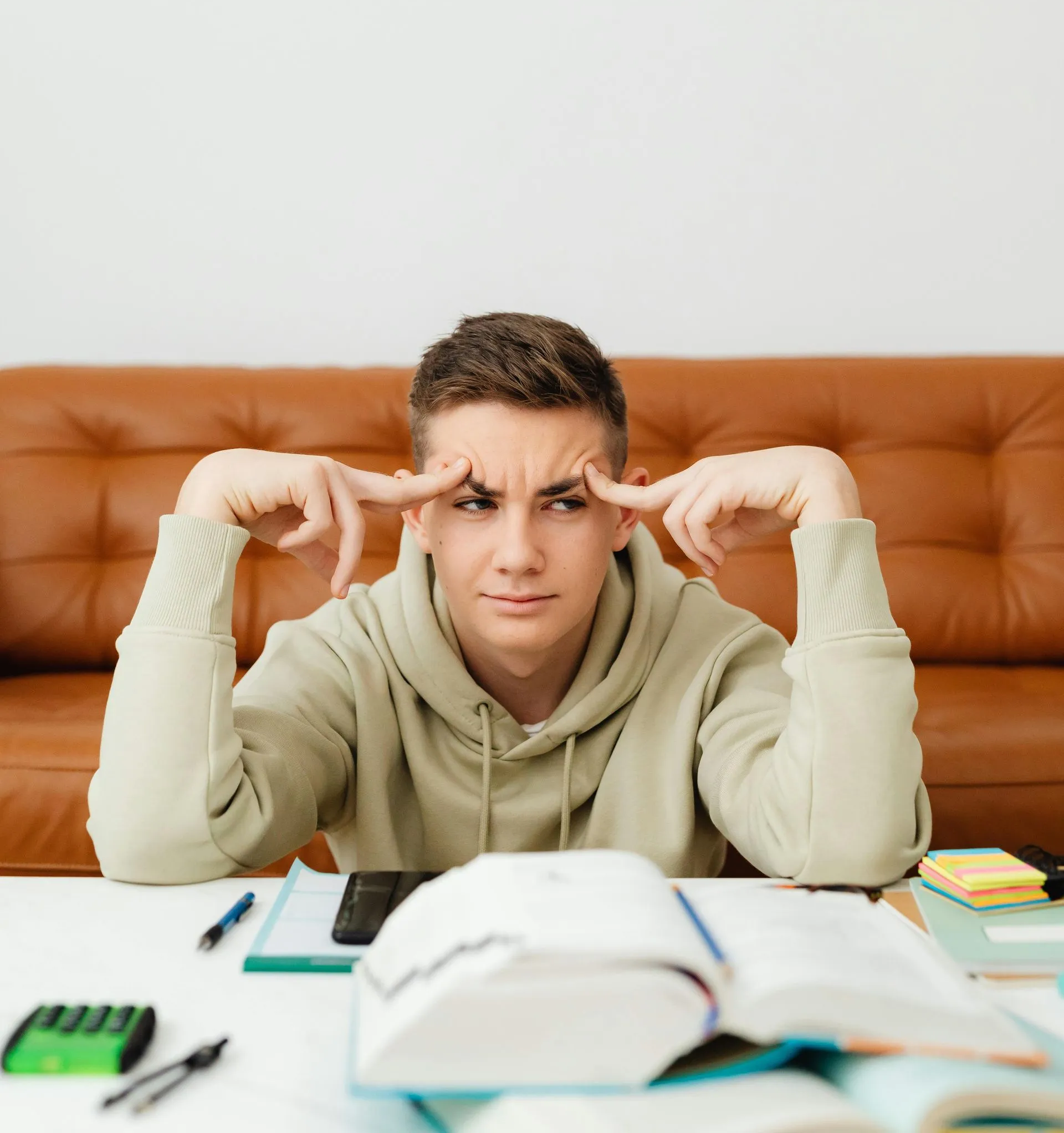 Teenager with furrowed brow touches temples, studying at a desk with books and calculator, indoors.
