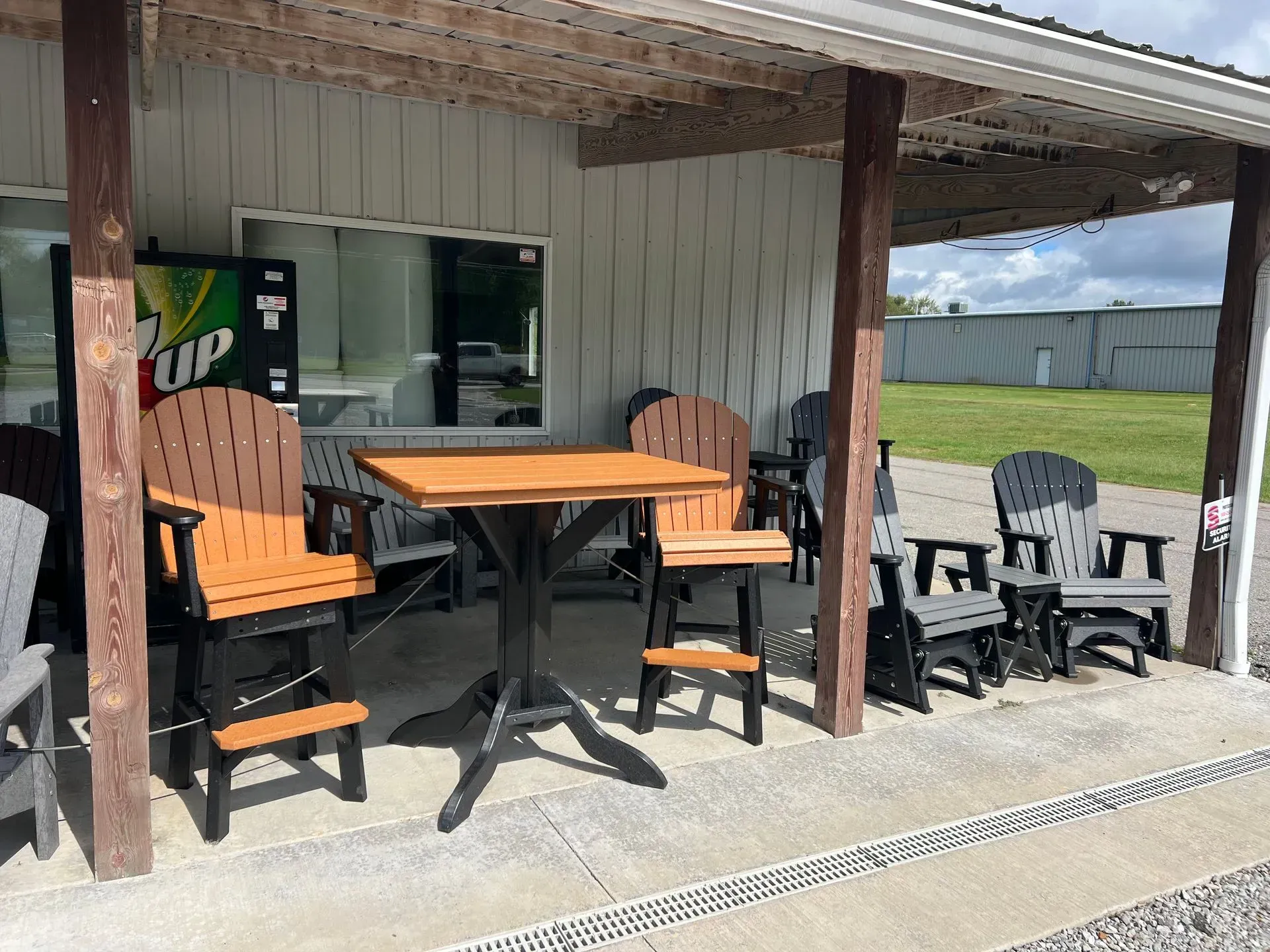 Outdoor furniture display: black and orange bar stools, table, and Adirondack chairs under a building's overhang.