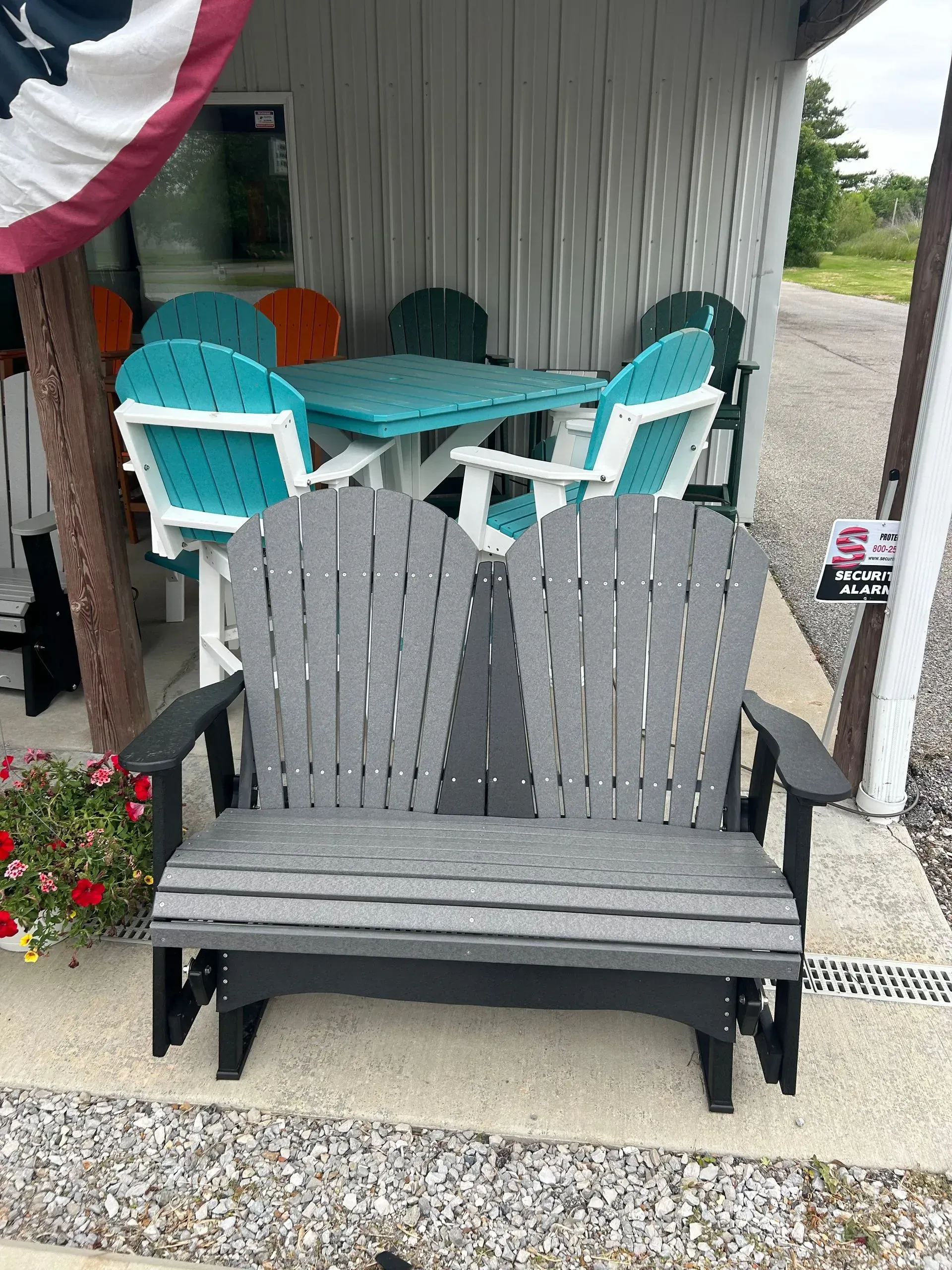 Outdoor furniture display. Gray double glider bench, teal table/chairs, colorful flags, storefront setting.