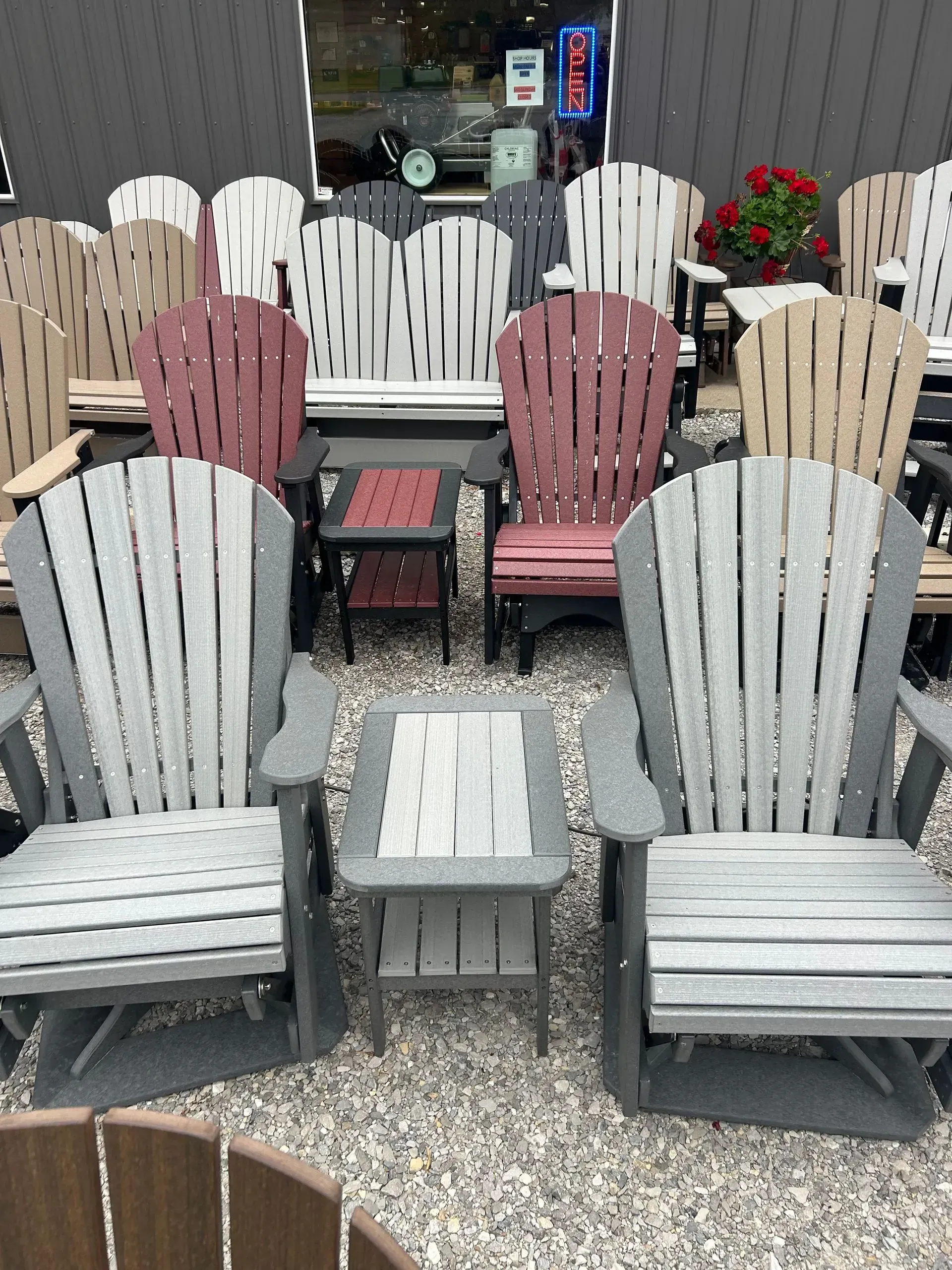 Outdoor furniture display: Adirondack chairs in various colors with small side tables, on a gravel surface.