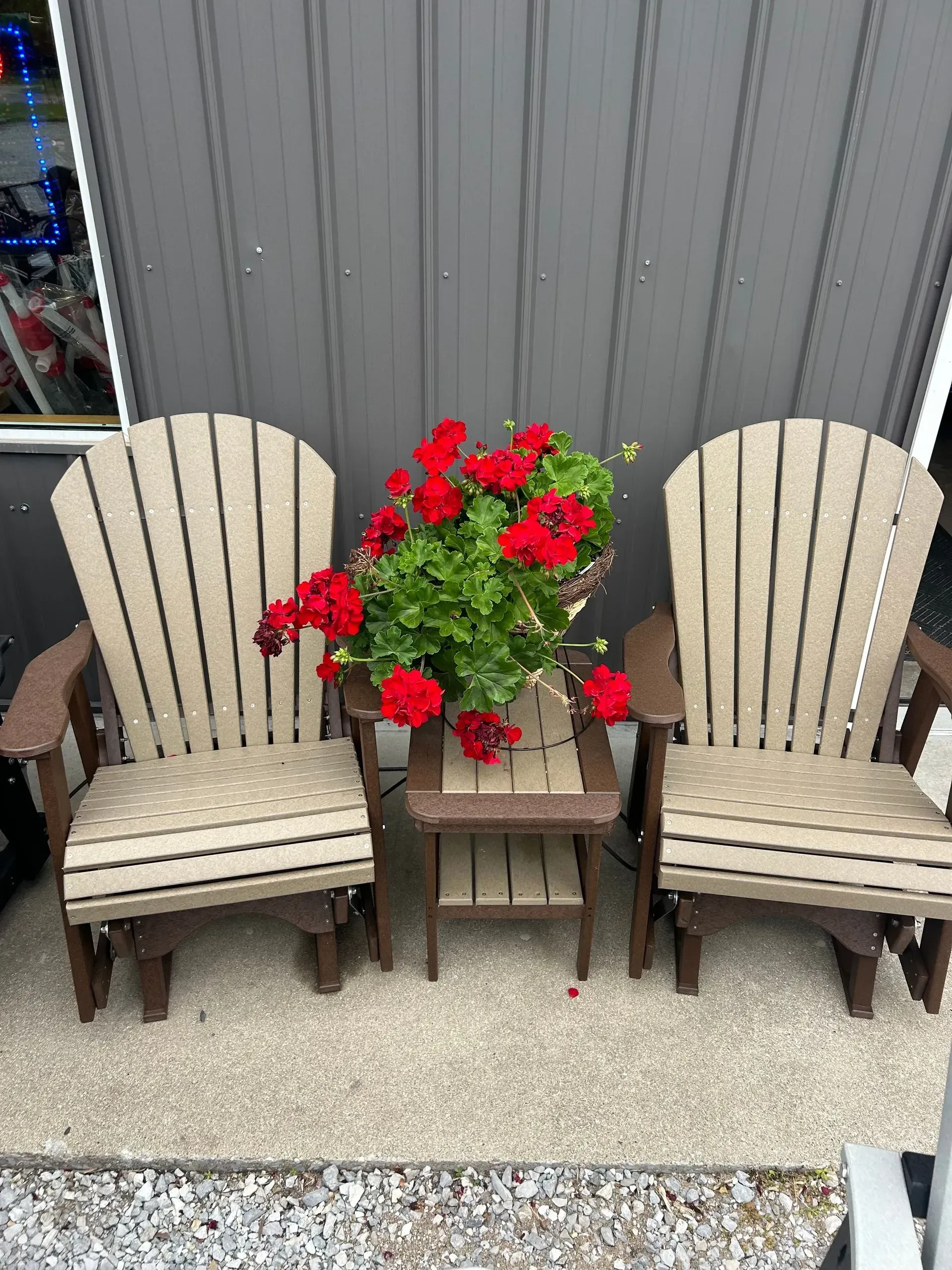 Two beige Adirondack chairs and small table with red flowers on a porch.