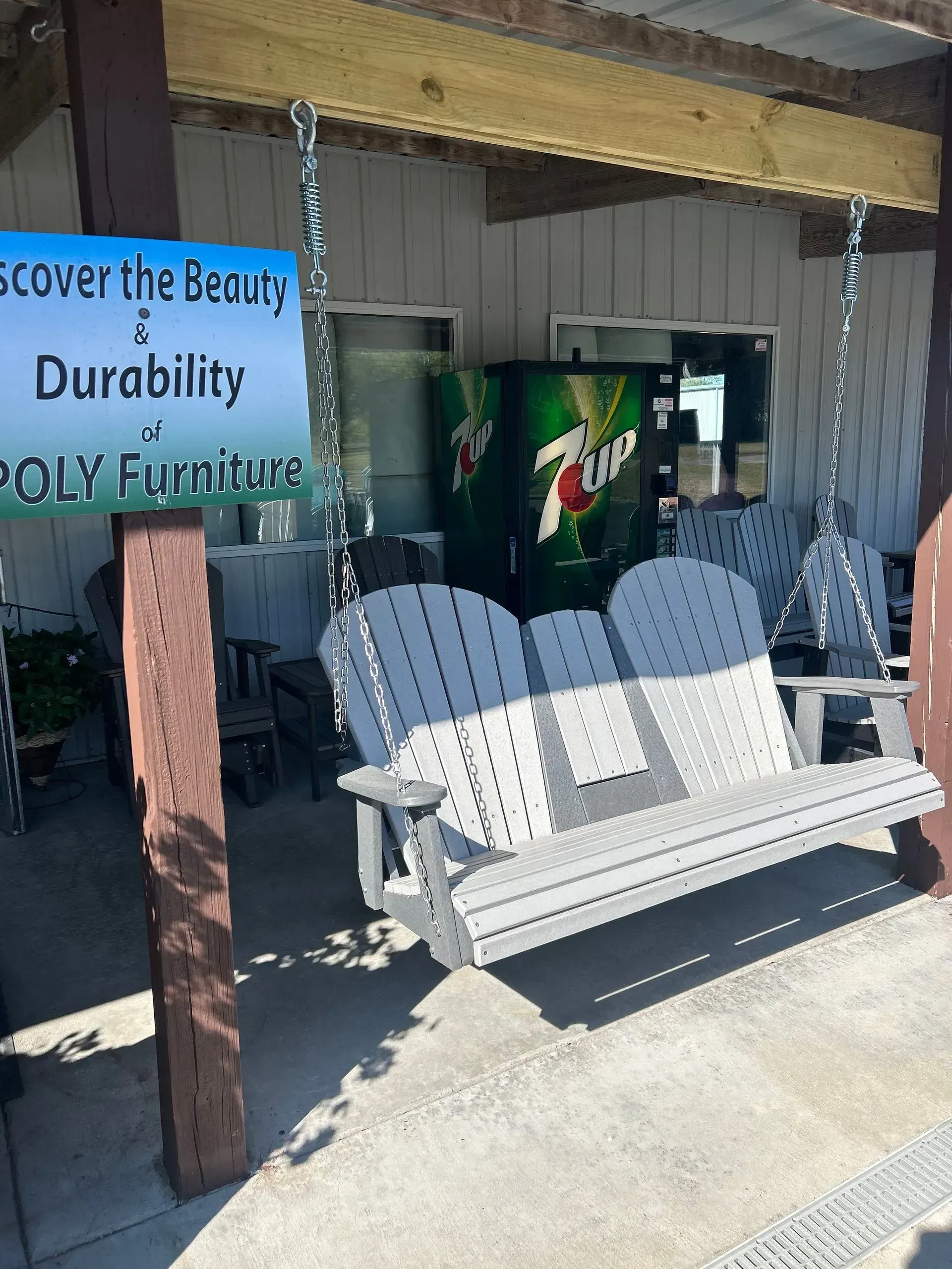 Outdoor display of poly furniture, including a gray swing, under a building overhang. Sign reads