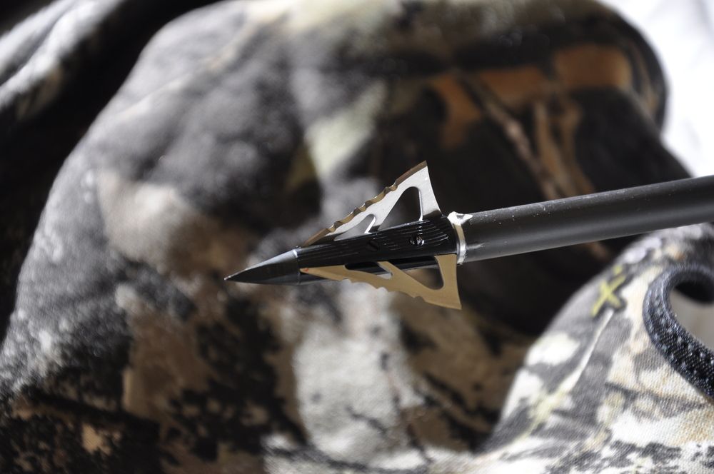 Black arrow with silver blades, pointed towards the camera, against a backdrop of camouflage fabric.