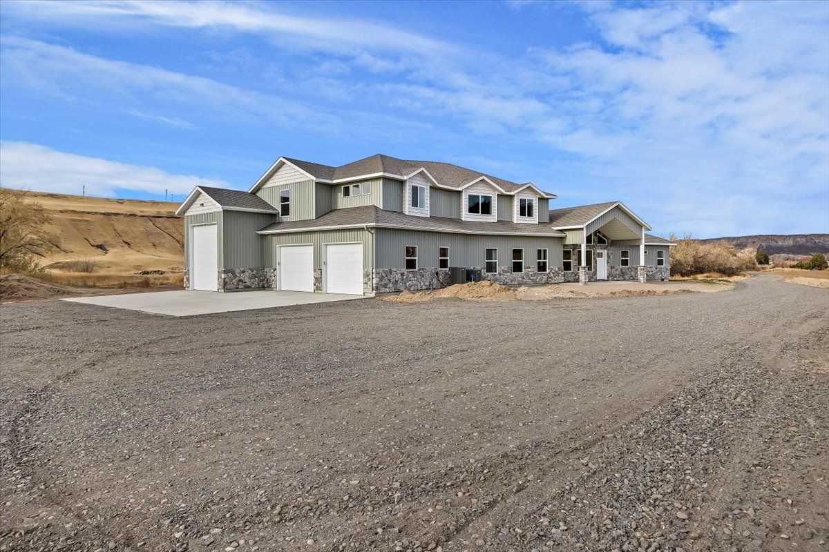 Large two-story house with attached garage, light blue siding, stone accents, gravel driveway, and a blue sky backdrop.