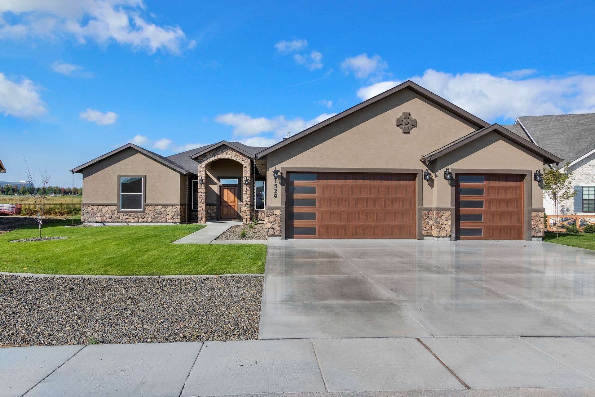 Tan stucco house with brown garage doors, stone accents, and a wide concrete driveway.