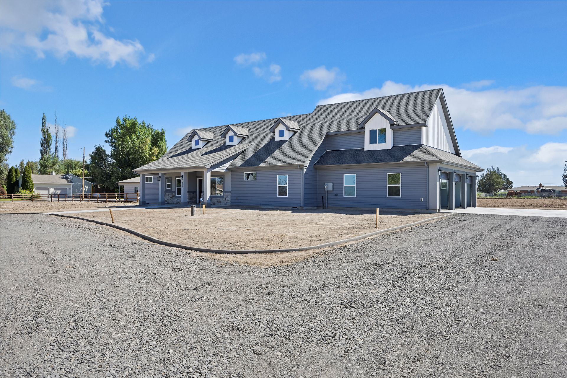 Blue house with gray roof, three dormers, and three-car garage on a gravel driveway under a blue sky.