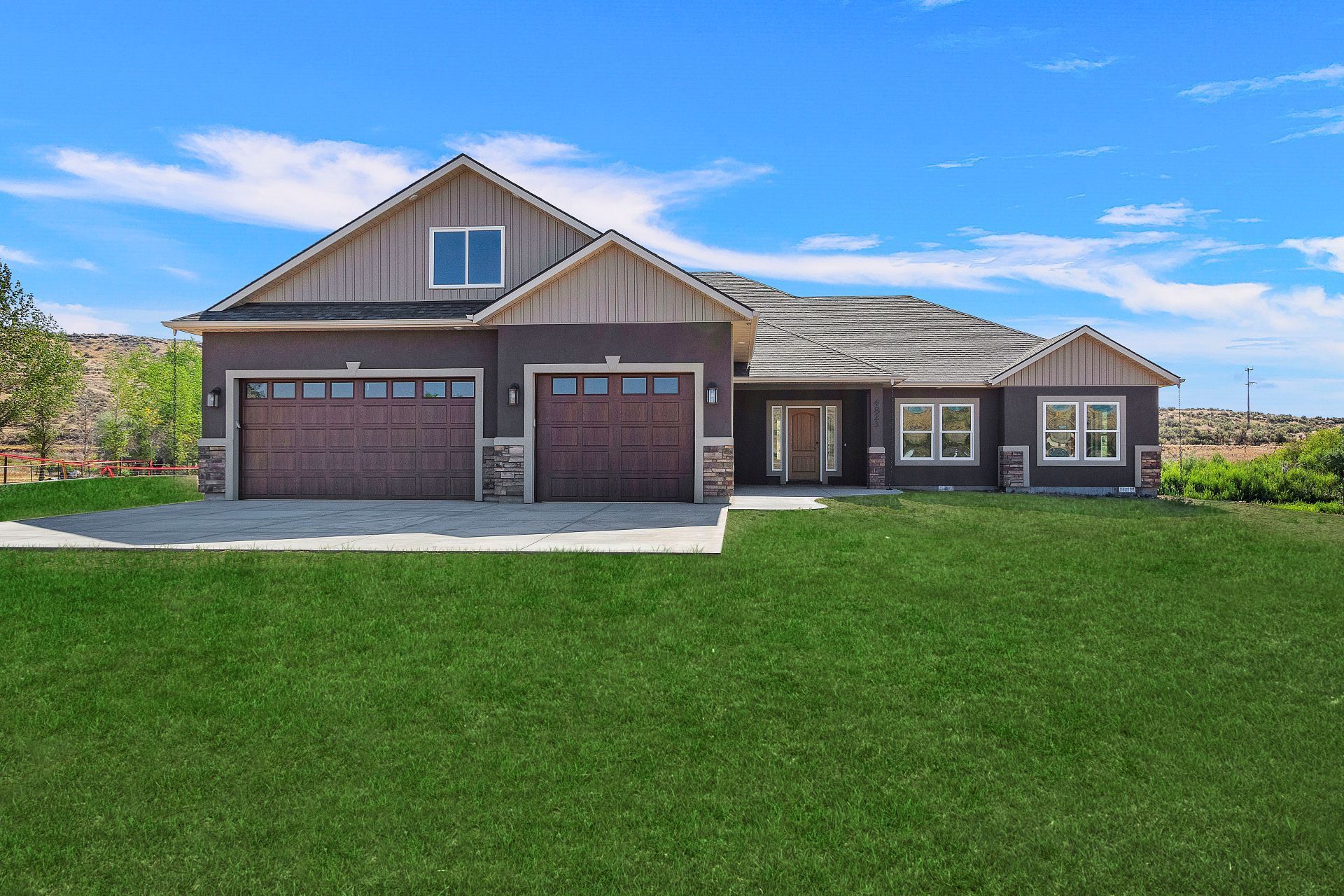 Brown house with two-car garage, front door, and green lawn under a blue sky.