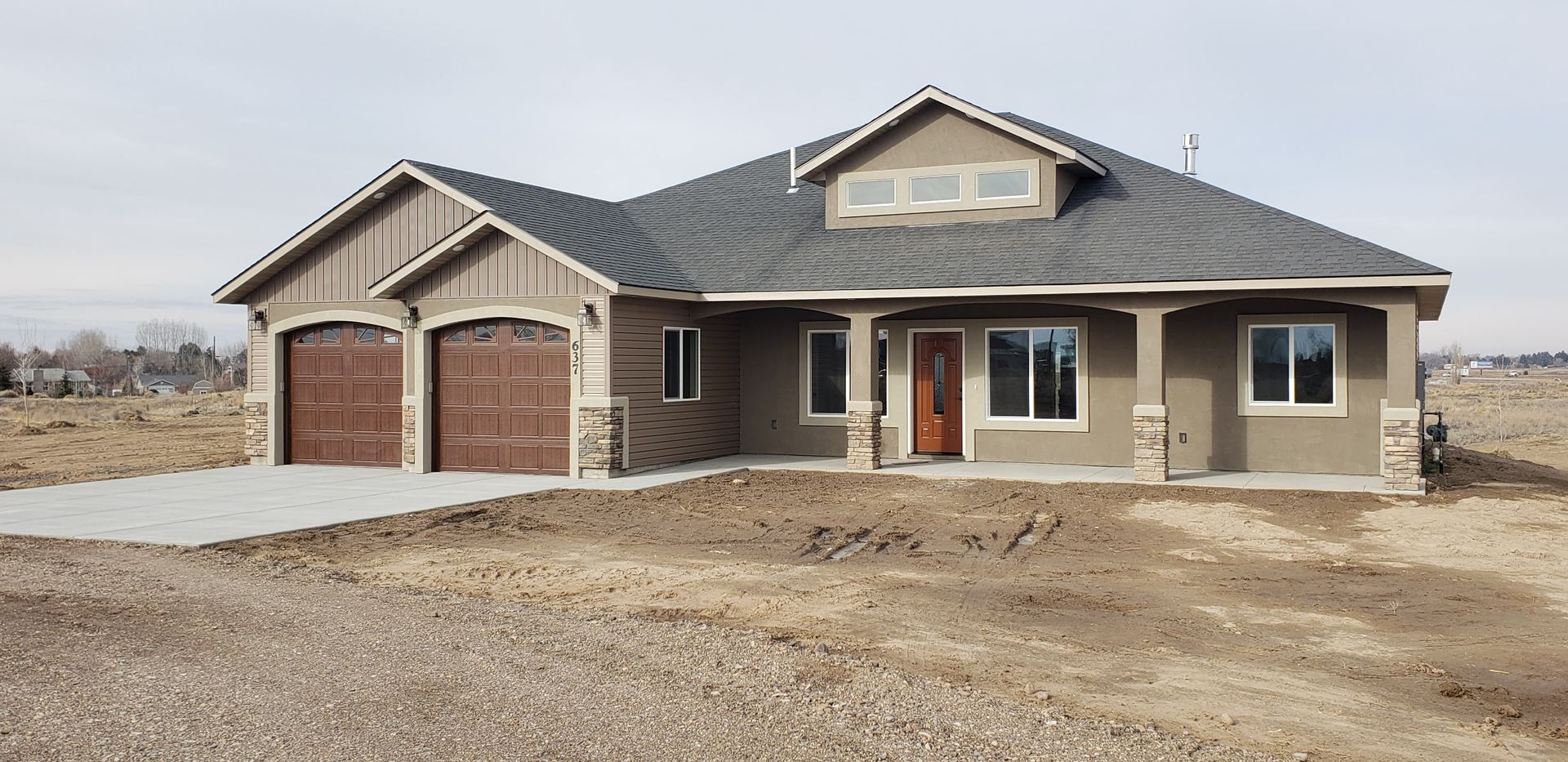A one-story house with a two-car garage is under construction on a dirt lot on a cloudy day.