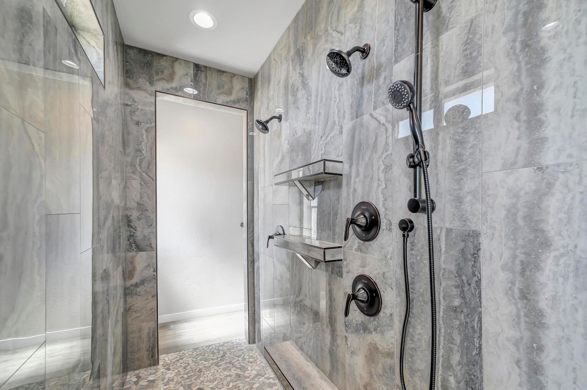 Modern bathroom with gray tiled walls, pebble floor, multiple shower heads, and glass door.