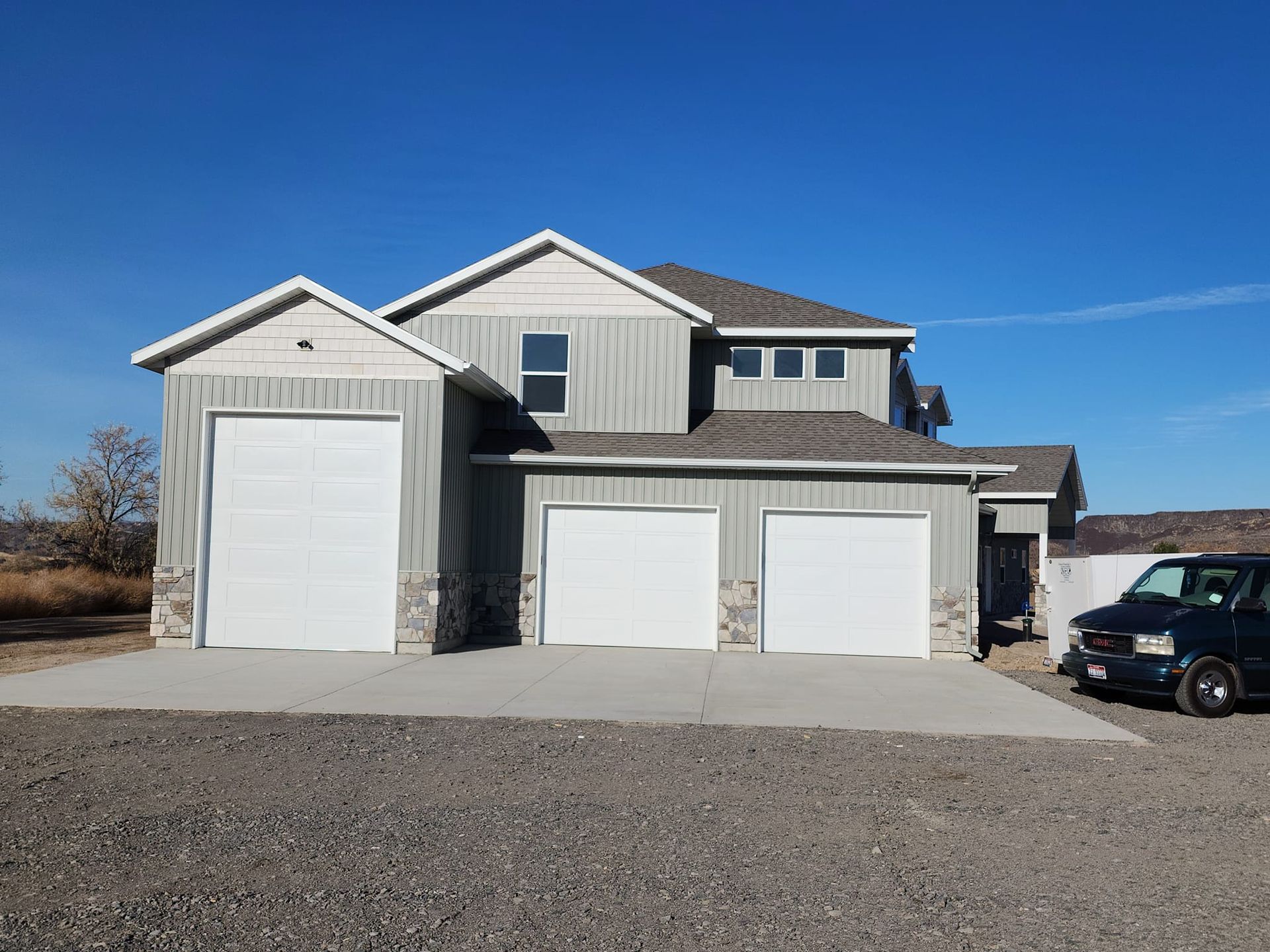 Three-car garage with a two-story house, gray siding, white garage doors, and a blue sky.