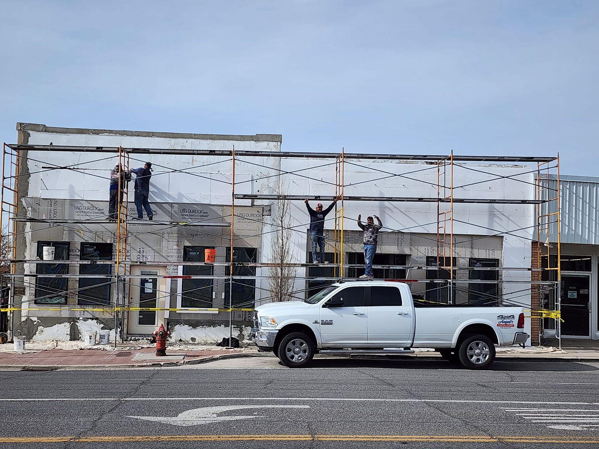 Construction workers on scaffolding, white pickup truck parked in front of building with exposed walls, work in progress.
