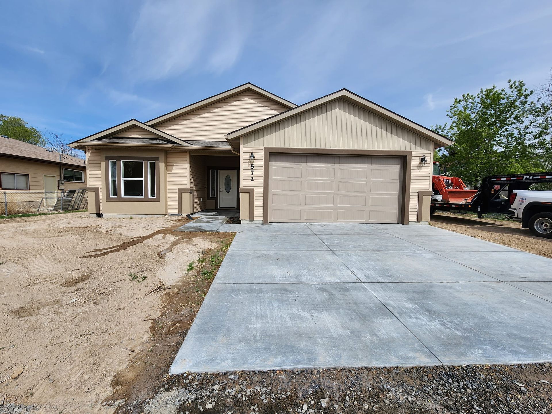 Tan-colored house with a concrete driveway and garage door. Construction vehicles parked to the right.