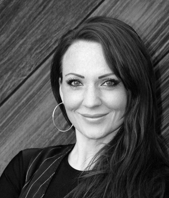 A black and white photo of a woman smiling in front of a wooden wall. Dana Miller