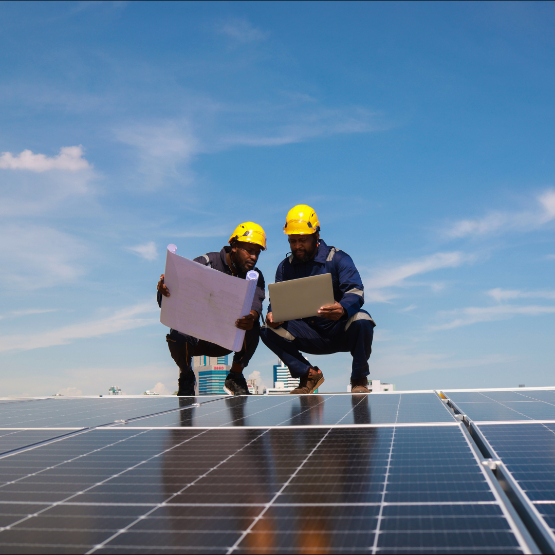 Two workers in hard hats examine a blueprint and laptop on a solar panel array against a blue sky.