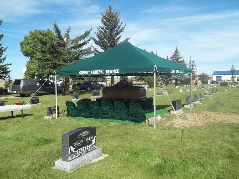 Funeral service outdoors; casket under green canopy, green chairs, grave markers in cemetery.