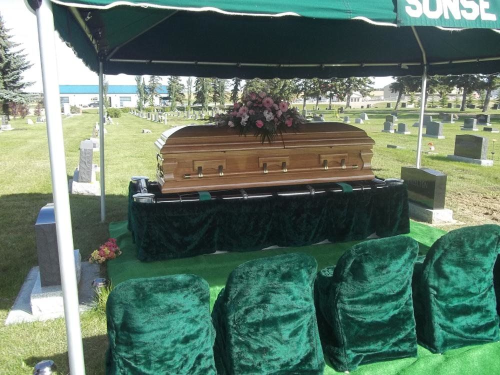 A casket draped in green velvet, topped with flowers, under a green canopy at a cemetery.