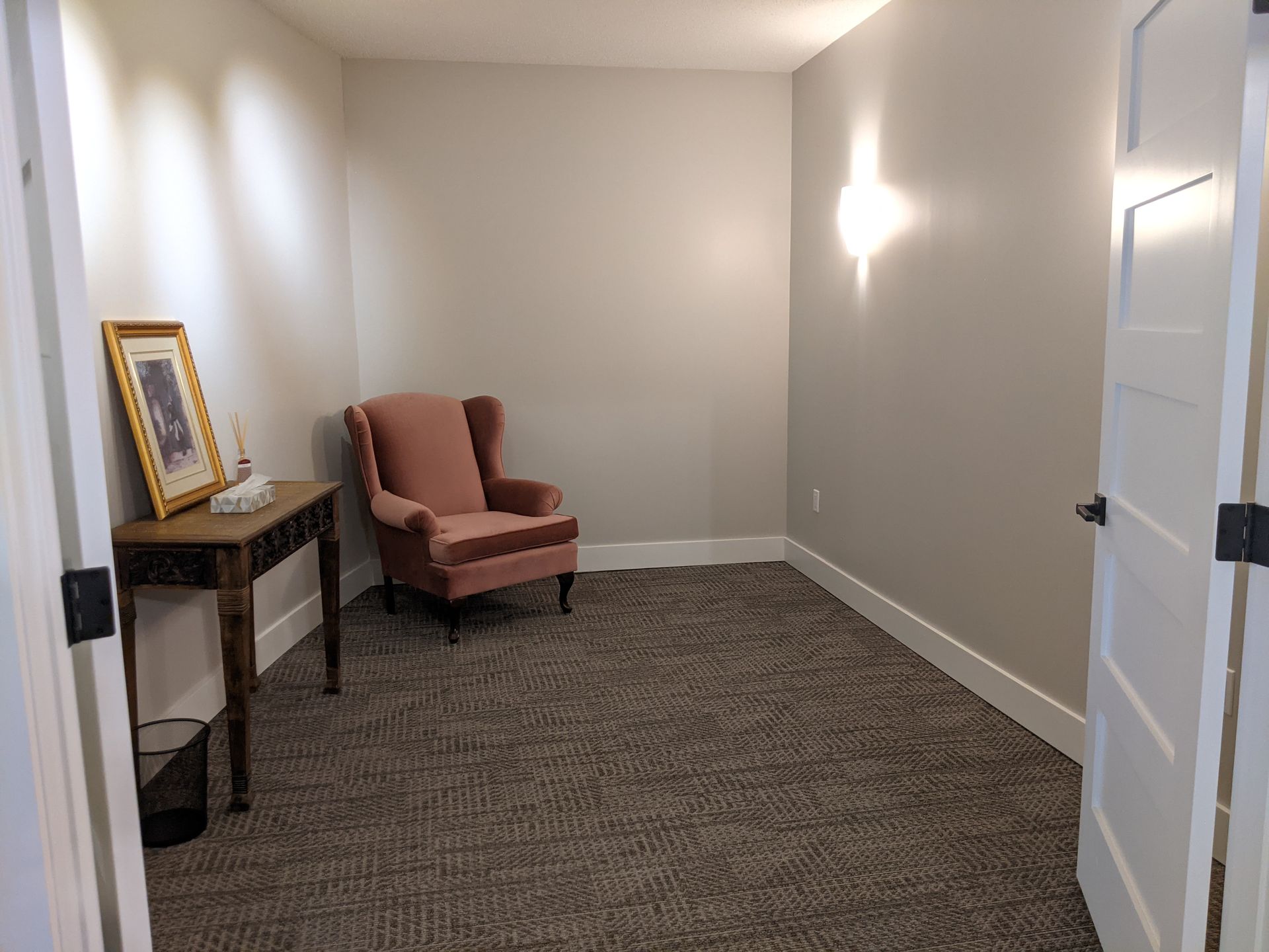Empty room with mauve chair, small table, and framed picture. Light-colored walls and carpet; door on the right.