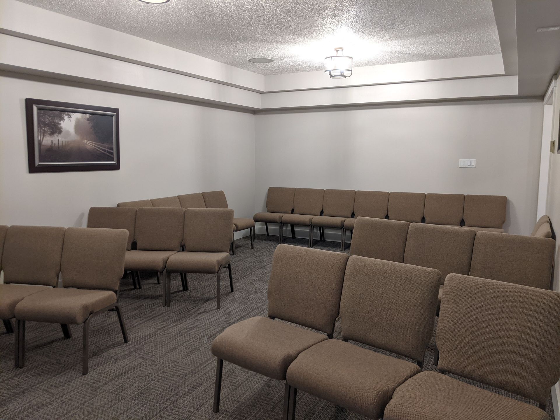 Empty room with rows of brown chairs, a framed landscape, and a textured ceiling.