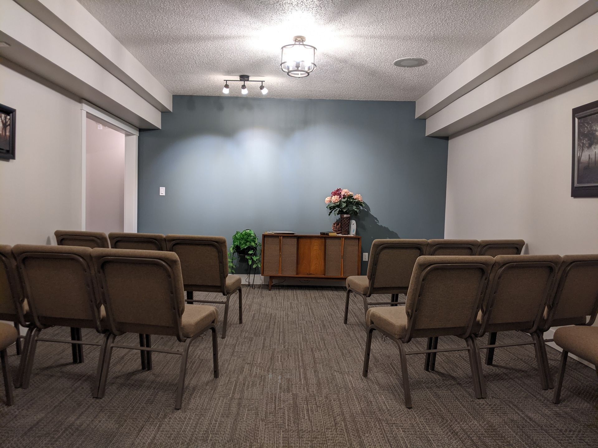 Rows of brown chairs face a wooden cabinet with flowers in a room; a blue wall backdrop.