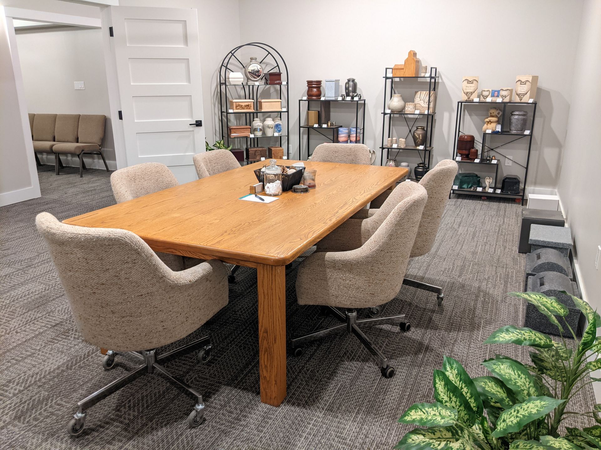 Meeting room with wooden table, six chairs, shelves with items, carpet, and a plant.