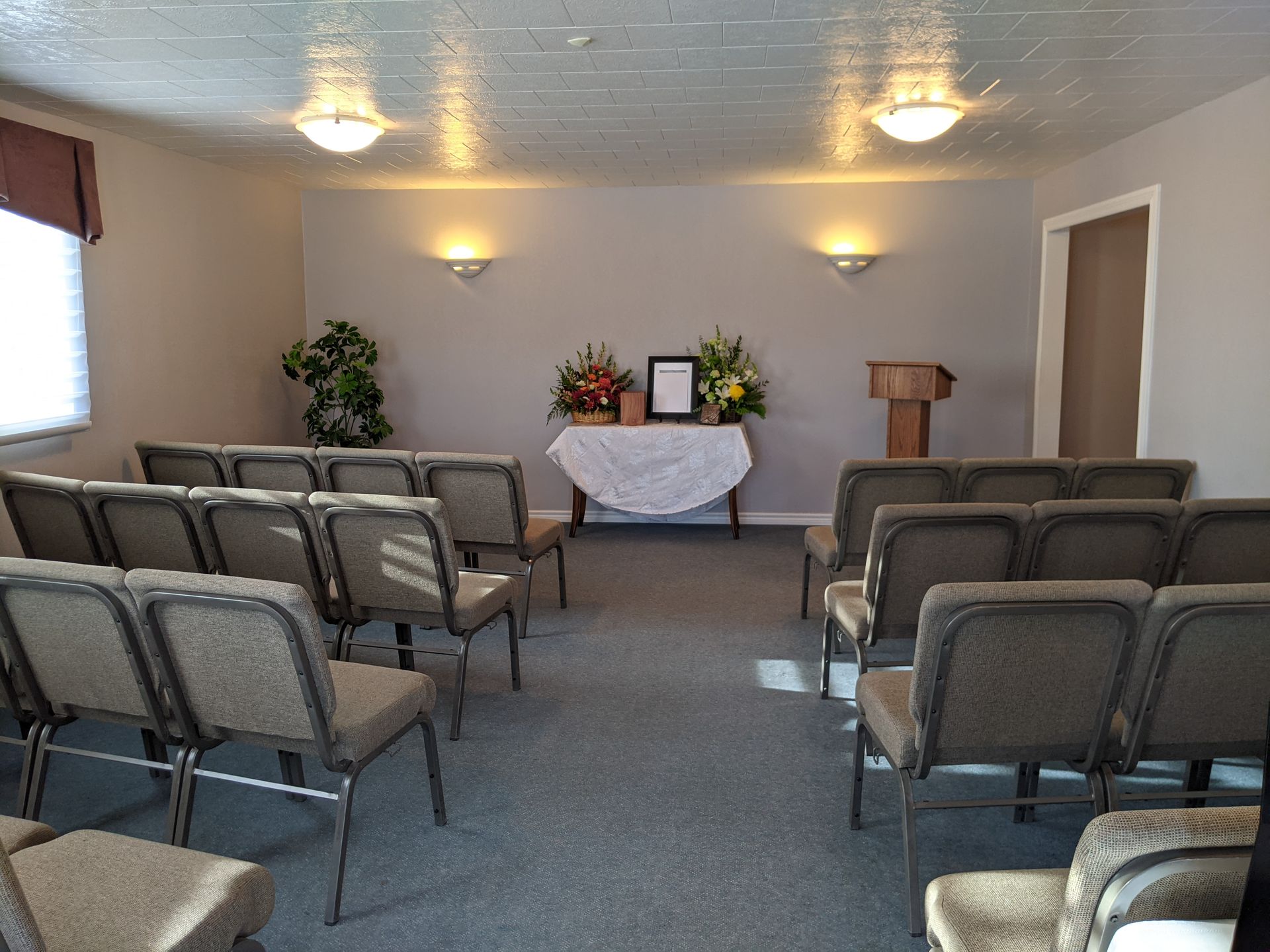A chapel with rows of chairs facing a table with flowers and a framed photo. A podium is to the right.