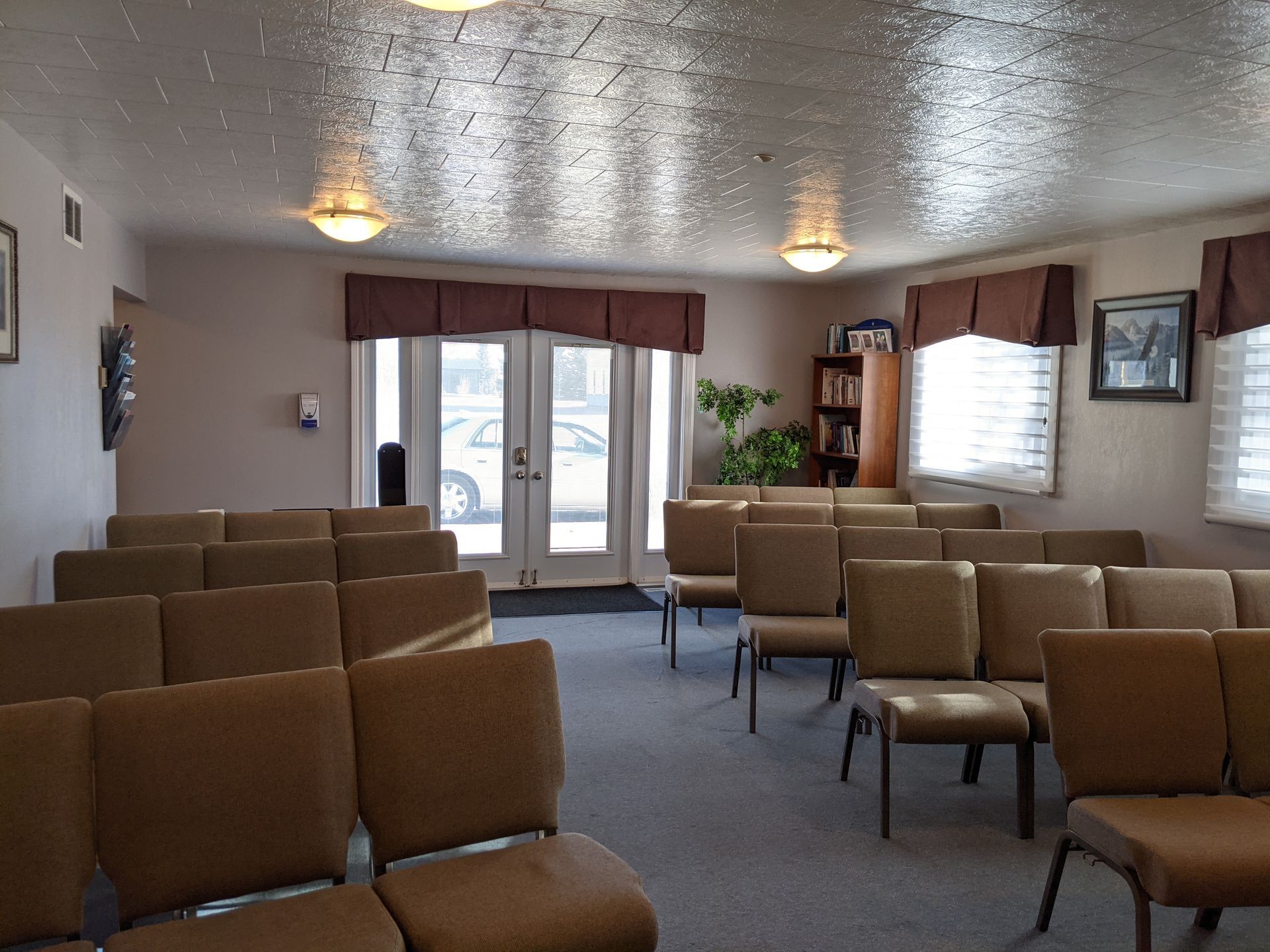 Interior view of a chapel with rows of chairs facing a set of double doors. Beige walls, carpet, and chairs.