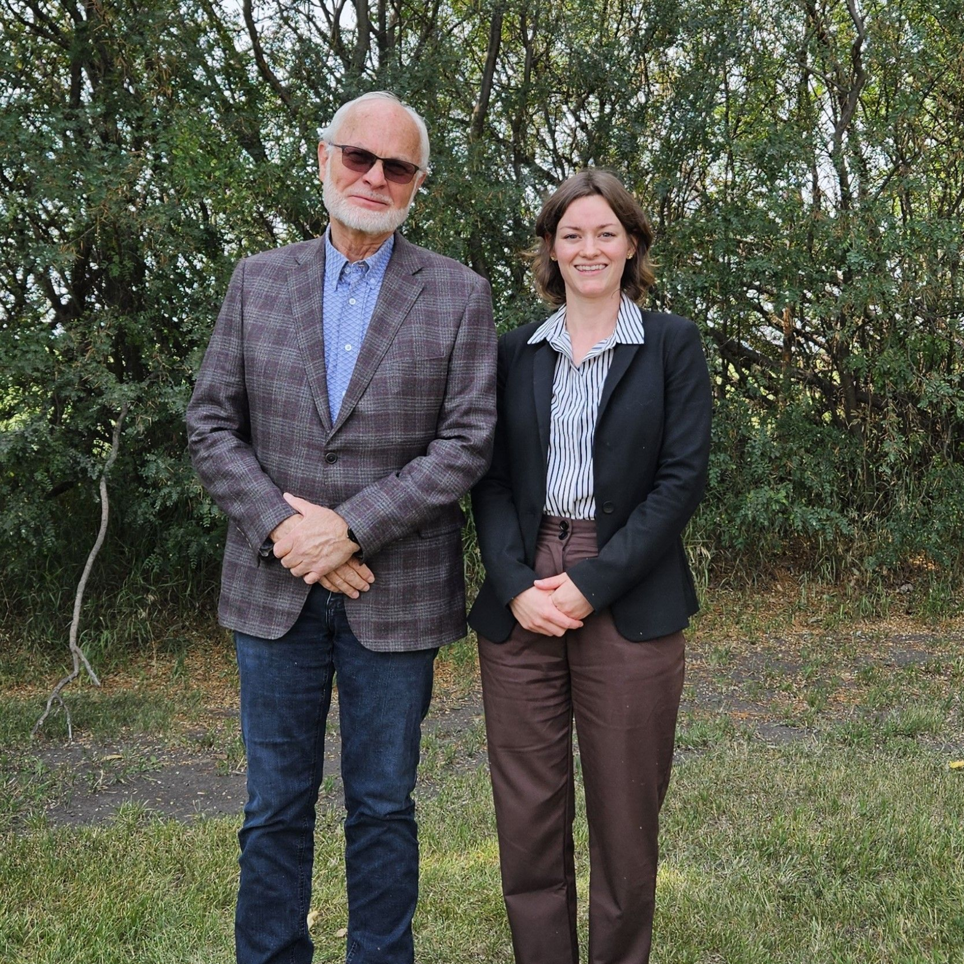 A man and woman stand outside. The man wears a blazer and sunglasses. The woman wears a blazer.