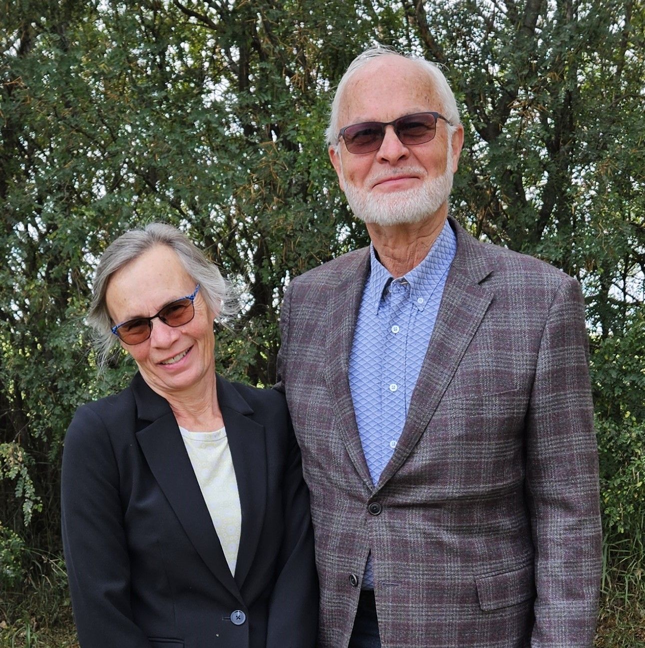 Man and woman in sunglasses standing in front of greenery. The man wears a blazer and the woman wears a jacket.