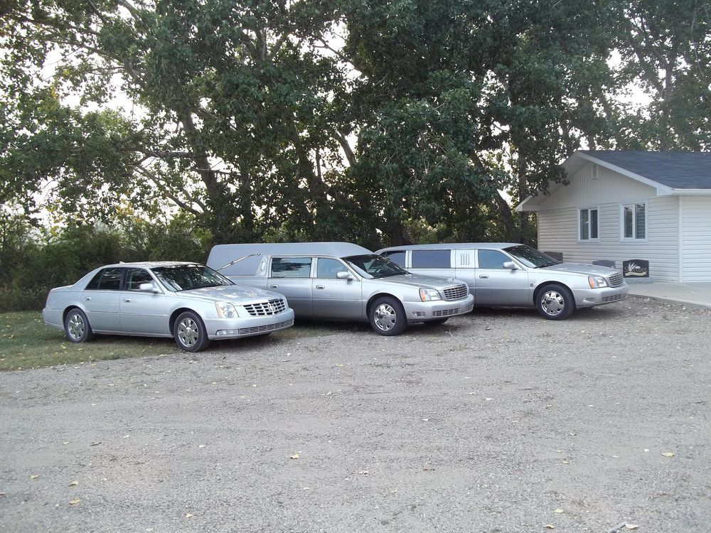 Three silver Cadillac vehicles parked near a building and trees. A hearse is in the middle.