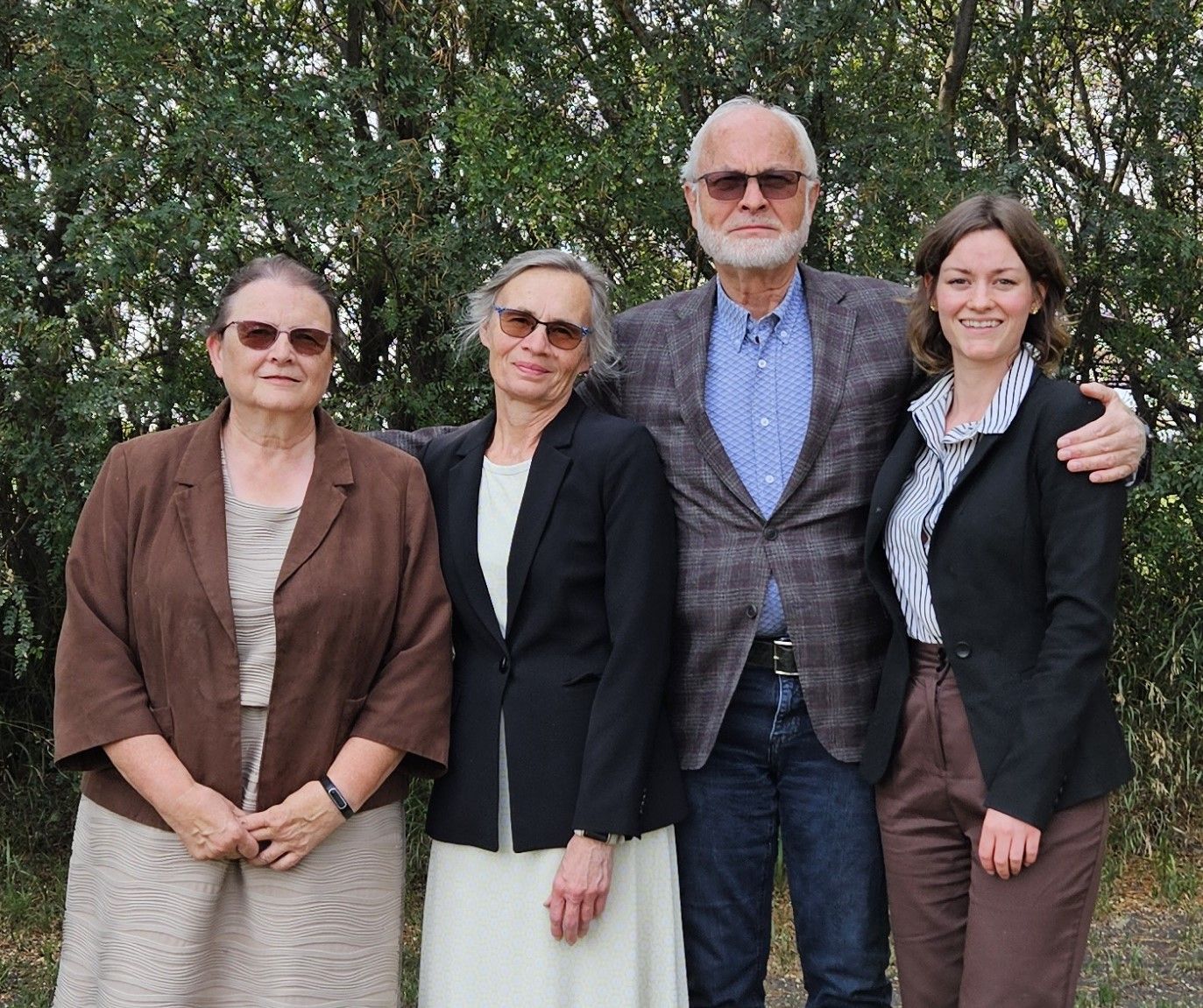 Four people standing together in front of greenery; two women on the ends, two in the middle, man with arms around others.