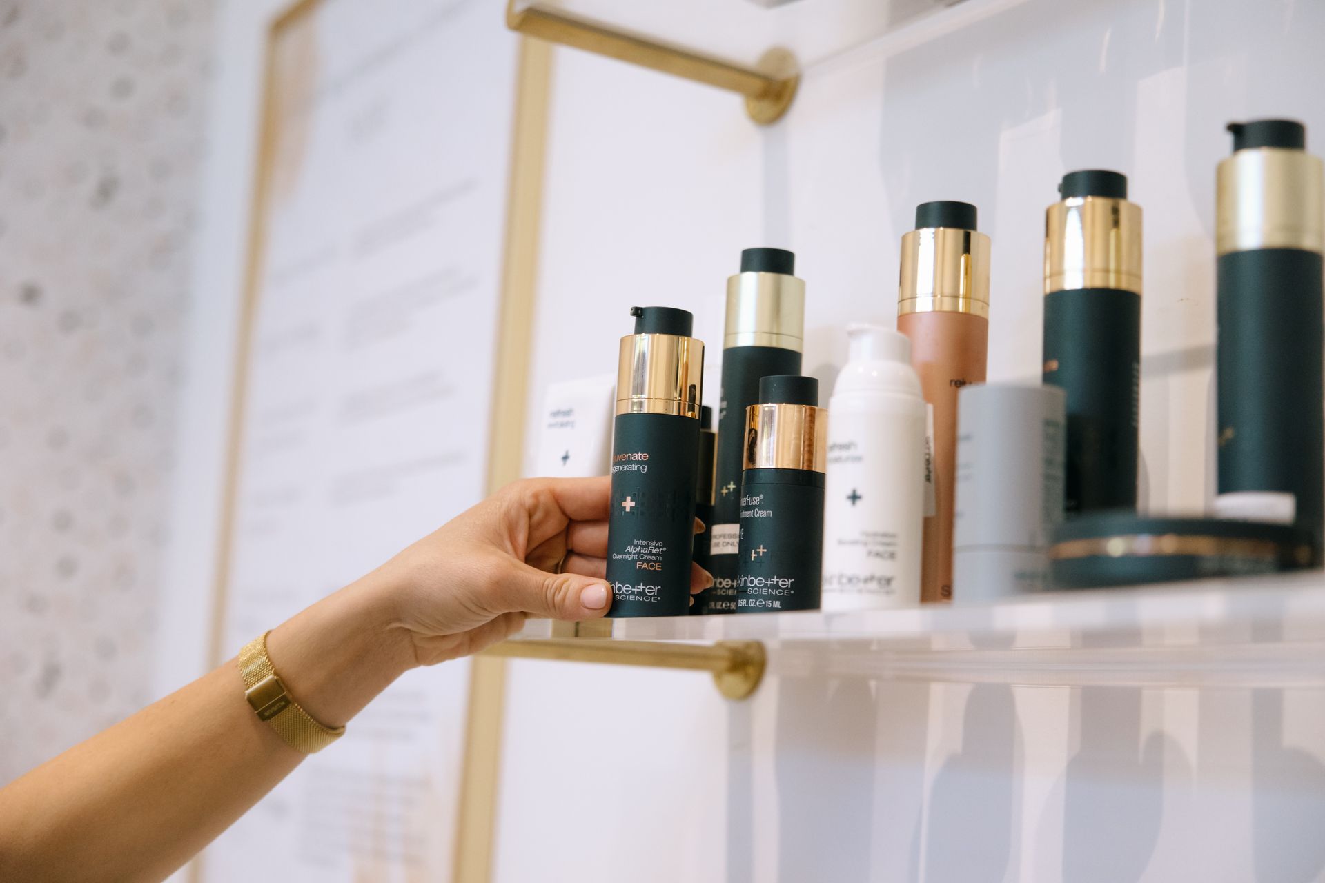 A hand reaching for a black and gold cosmetic bottle on a shelf with other products.