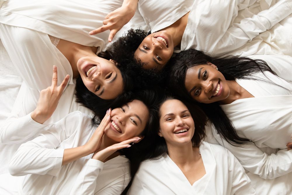 Five smiling women lying on a bed in white pajamas.