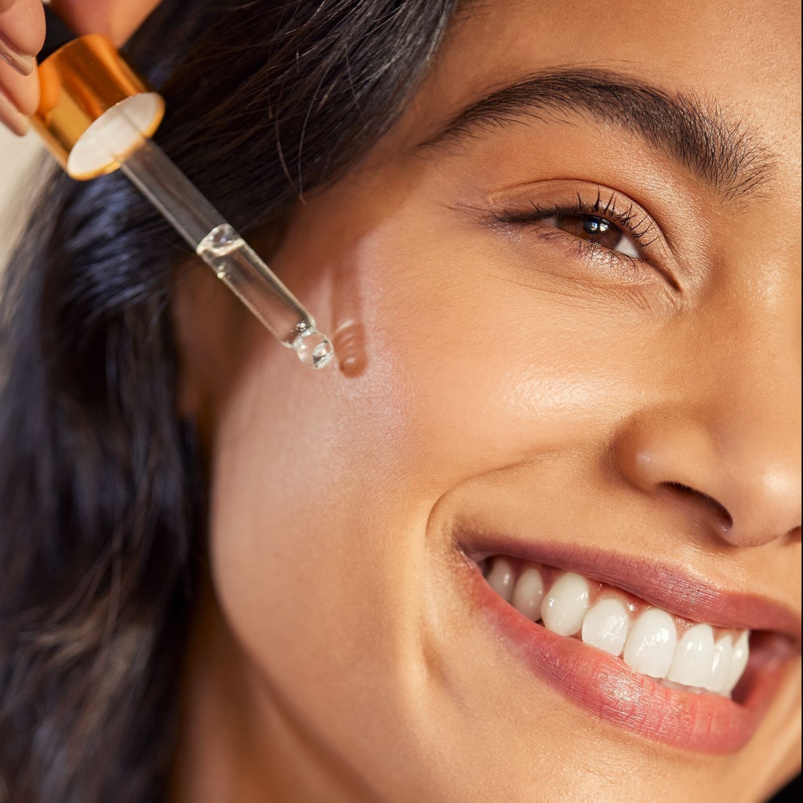 A woman is smiling while applying a serum to her face.