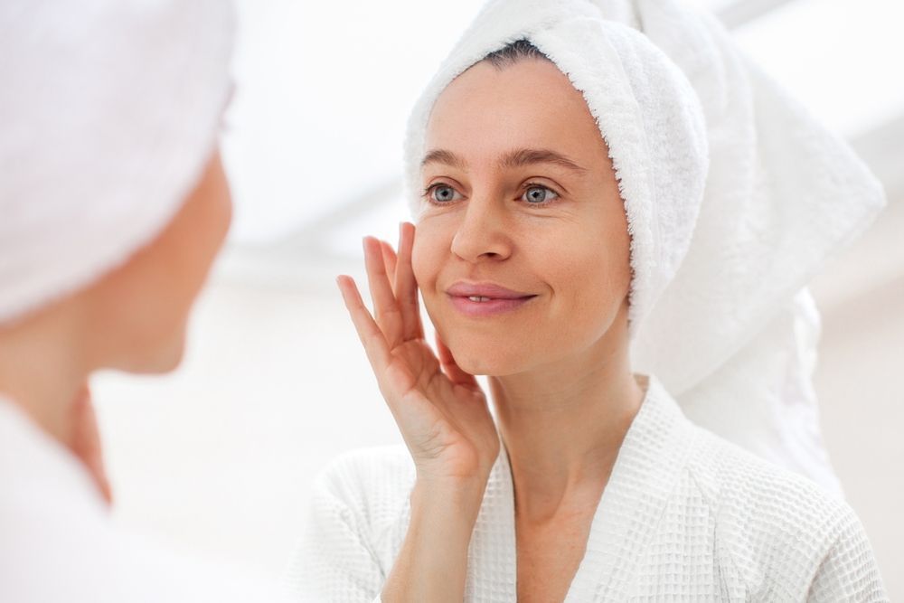 Woman in robe and towel looking at mirror, touching her face. Bright, neutral background.